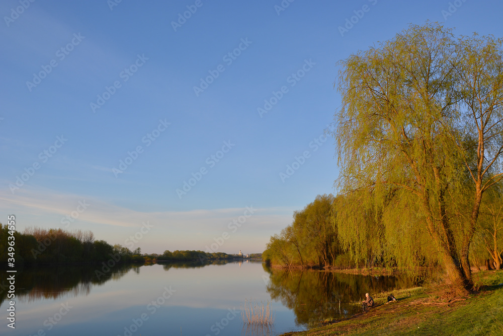 Summer landscape with a river and trees on and a fisherman in the foreground and the St. George Monastery on the horizon. Veliky Novgorod. Russia.