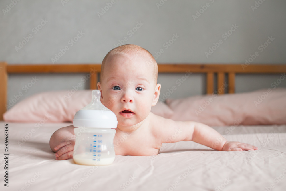 Artificial feeding. The baby lies on the bed on her tummy and looks at the feeding bottle in front of him with the adapted milk formula Artificial feeding. The baby lies on the bed on her tummy and looks at the feeding bottle in front of him with the adapted milk formula