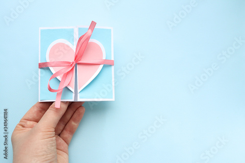 Fototapeta Naklejka Na Ścianę i Meble -  A small blue holiday card with a pink heart and a ribbon in the hand of a Caucasian man on a blue background. Space for text.Selective focus. Valentine's day and Mother's day greeting cards.copy space