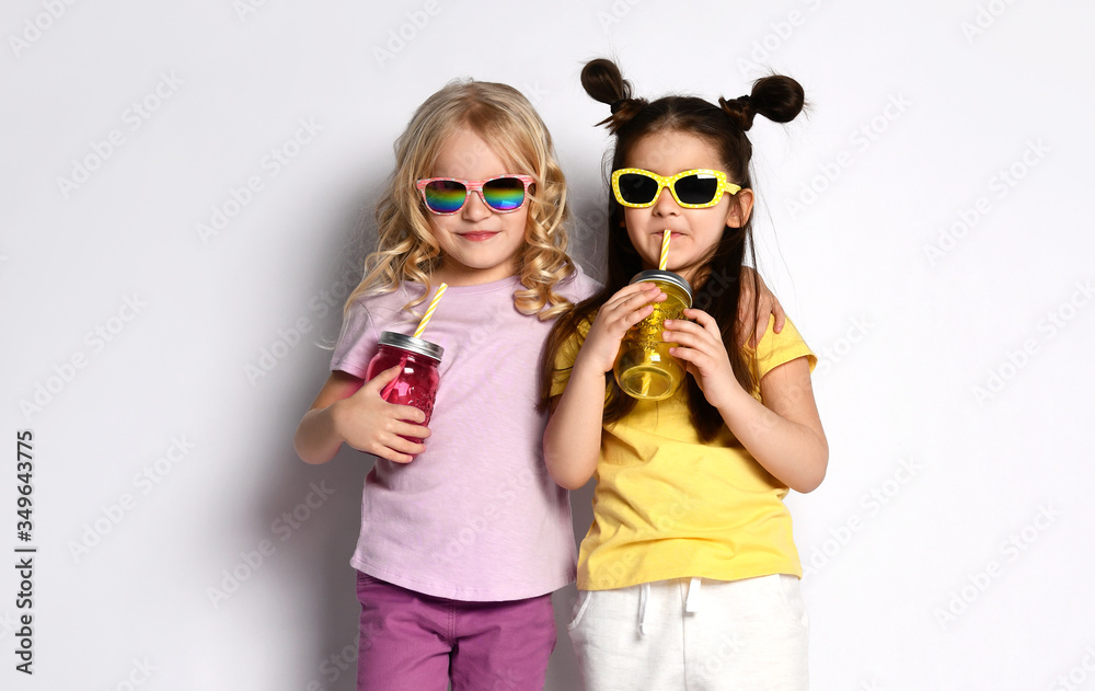 Two little models in sunglasses, colorful casual clothes. Holding cocktail bottles, showing tongues, posing isolated on white