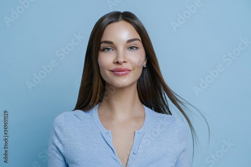 Indoor portrait of pretty woman feels happy and carefree during day off, has long straight hair, wears blue jumper, enjoys positive conversation, stands indoor. Women, lifestyle and well being concept © VK Studio