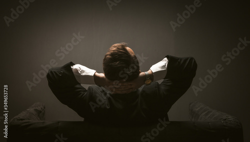 Businessman pondering new startup project sitting on a sofa. Rear view of Man in black suit sitting at couch in dark room while thinking with hands behind his head