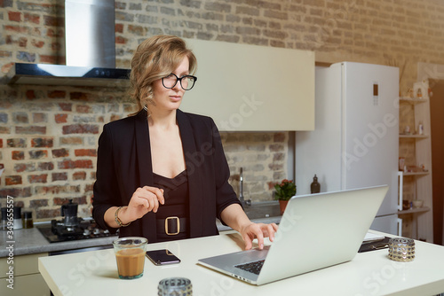A young woman in glasses works remotely on a laptop in her kitchen. A serious girl calmly browsing news on the internet at home. A lady preparing for a lecture on a video call..