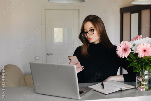 Young professional businesswoman tired while using smartphone and messaging at work on her project with laptop in comfortable home office with flowers. Remote work concept.