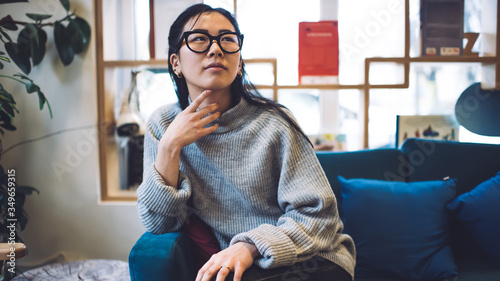 Fotografi Asian female in comfortable knitwear sitting on sofa with dreamy look