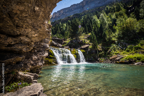 Waterfall in crystal clear river with mountains and forest on sunny summer day