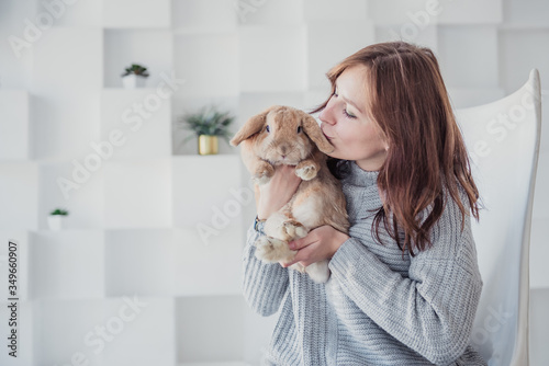 A young woman in a gray sweater is sitting at home in isolation with a rabbit on a light background © Ekaterina Voitenkova