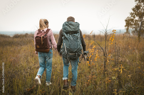 Hiking in autumn nature, couple of backpackers makes their way across the field, rear view of man and woman with backpacks and hiking poles walking in nature outdoors. Hiking concept