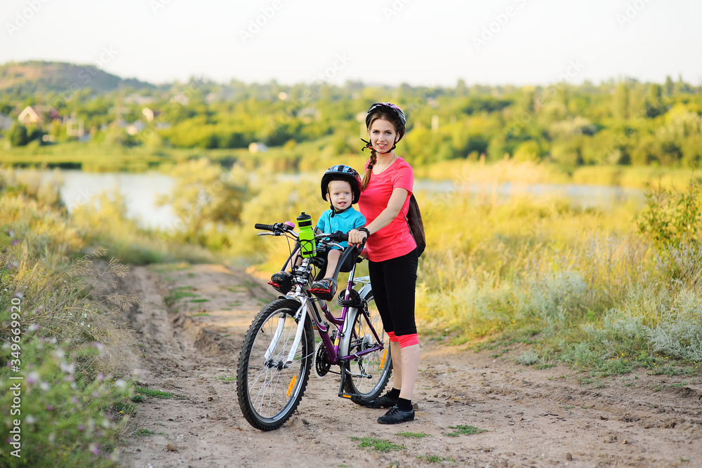 a small child in a protective helmet smiles sitting on a child's Bicycle seat on a large mountain bike with his mother on the background of green grass and nature. Active lifestyle, sports, childhood