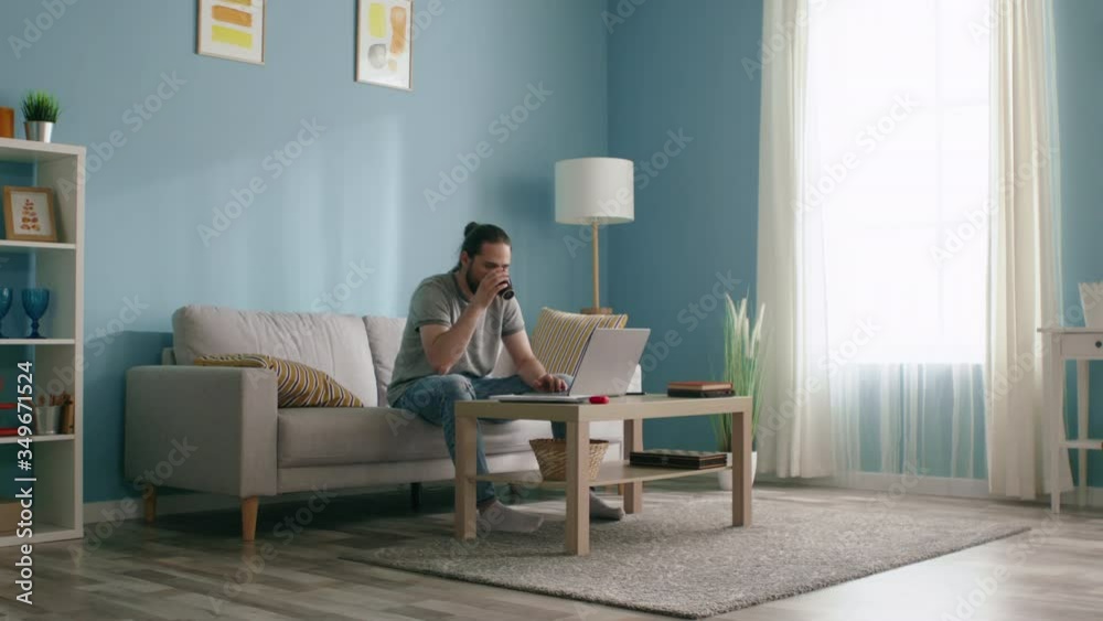Young man, freelancer, is working at laptop in living room, drinking coffee, enjoying freelance lifestyle, focusing on daily tasks, Slow motion. Young man, freelancer, is working at laptop in living room, drinking coffee, enjoying freelance lifestyle, focusing on daily tasks, Slow motion.