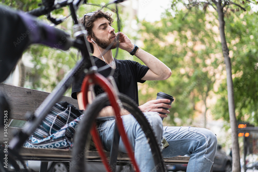 Obraz premium Hipster guy relaxing on a bench in the street drinking an American coffee in the city and listening to music with earphones.