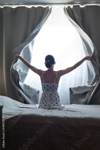 Woman alone at home , opens the curtains letting in bright ray of sunlight, concept of brighter future. To meet bright day.