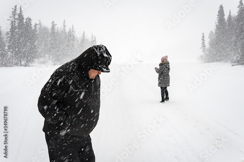 Two people standing on a snow field during a snowstorm next to Glenn Highway, on the way to Anchorage, in Alaska. They are a man in front with a black jacket and a woman in the back with gray coat.