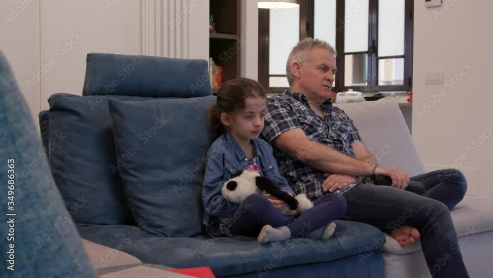 Movie at home with divorced dad and daughter. Unhappy family with man and female child watching television and sitting on sofa at home, snorting while watching boring show or film on TV Movie at home with divorced dad and daughter. Unhappy family with man and female child watching television and sitting on sofa at home, snorting while watching boring show or film on TV