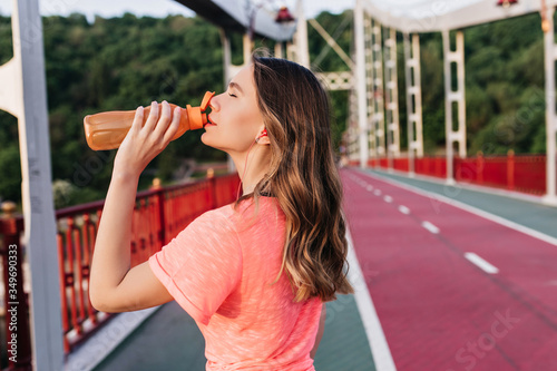 Pretty girl with wavy hair drinking water after maraphon. Refined caucasian lady posing at cinder track during training. photo