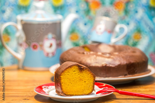 slice of carrot cake with chocolate sauce with teapot and cup of tea on blurred patterned background