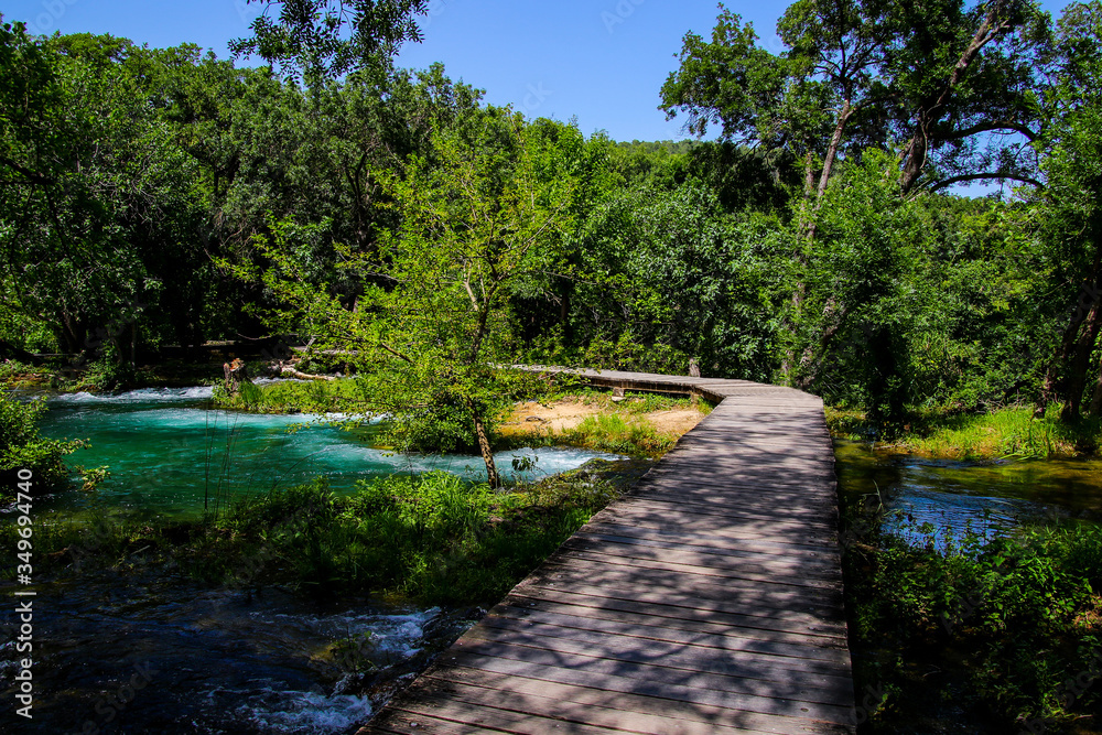 Fototapeta premium Wooden path built over the streaming waters of Krka National Park in Croatia - Tourists hiking in the forest along the river down to the waterfall in summer