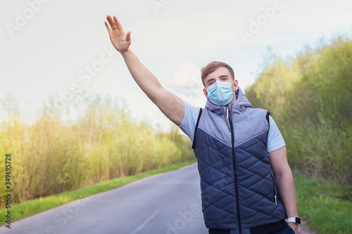 Young man, guy in medical mask for prevention of coronavirus, traveler trying to stop auto, car. Sad guy thand up gesture at road. Travel, journey, hitchhiking concept. Passenger. photo