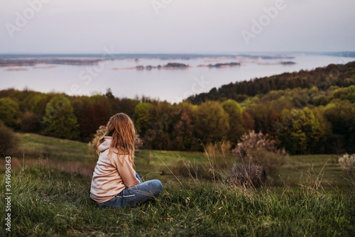  A girl with blond hair sits on the grass and looks into the distance in a pink raglan with a hood. Sunset. Time for reflection. Inner peace. Psychological relaxation