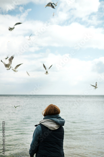 A lonely girl in a tank top and a pink sweater with a hood feeds seagulls by the sea. Blue sky with clouds and seagulls fly over the water. Peace in life. Internal conflict. Looking forward, back view