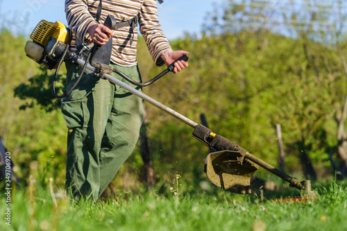 Midsection of unknown Young caucasian man farmer gardener standing in the field with string trimmer petrol Brushcutter ready to cut weed grass working on the farm cutting in the field in sunny day photo