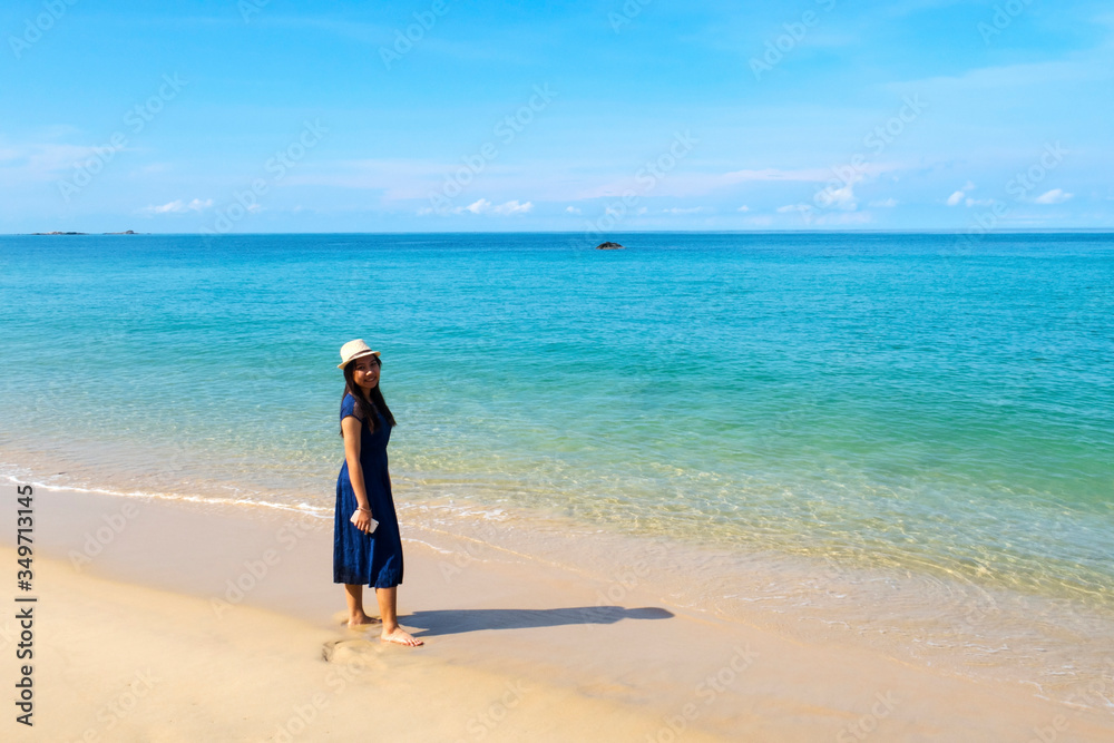 Cute Asian young teenager girl wearing sun hat & navy blue dress holding mobile phone or smartphone enjoying & admiring of deep blue sea view with soft calm waves touching tropical summer sand beach