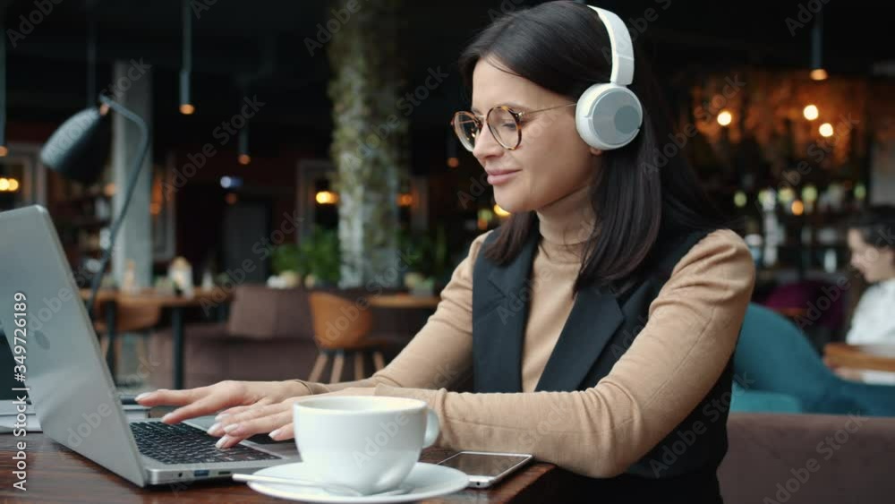 Happy businesslady is using laptop in cafe typing working online and listening to music through wireless headphones. Fun and business concept.