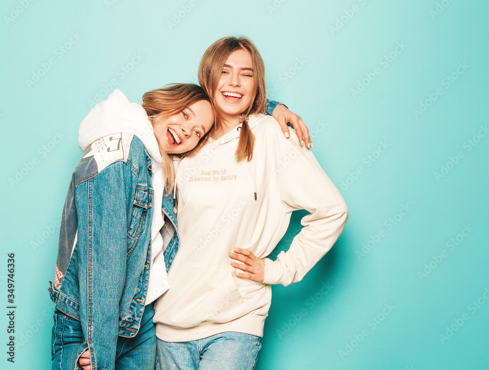 Two young beautiful brunette smiling hipster girls in trendy summer hoodie and jeans jacket clothes. Sexy carefree women posing near blue wall. Trendy and positive models having fun Two young beautiful brunette smiling hipster girls in trendy summer hoodie and jeans jacket clothes. Sexy carefree women posing near blue wall. Trendy and positive models having fun