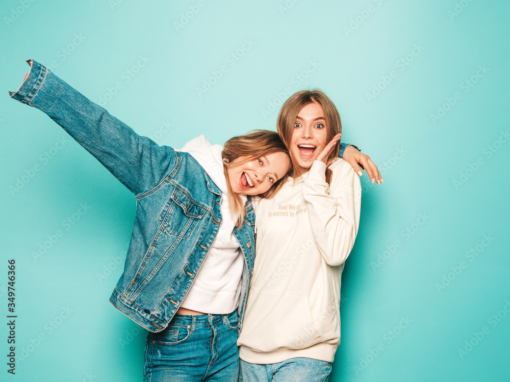 Two young beautiful brunette smiling hipster girls in trendy summer hoodie and jeans jacket clothes. Sexy carefree women posing near blue wall. Trendy and positive models having fun Two young beautiful brunette smiling hipster girls in trendy summer hoodie and jeans jacket clothes. Sexy carefree women posing near blue wall. Trendy and positive models having fun