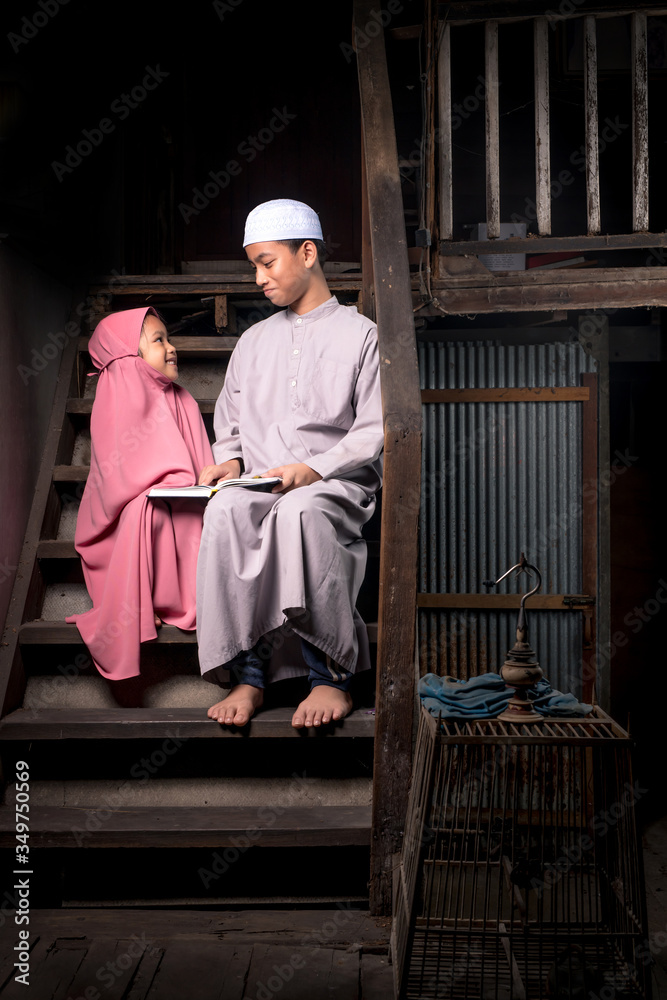 A brother and a cute little girl, an Asian Muslim, is practicing reading Quran with faith on the stairs in an old wooden house.
