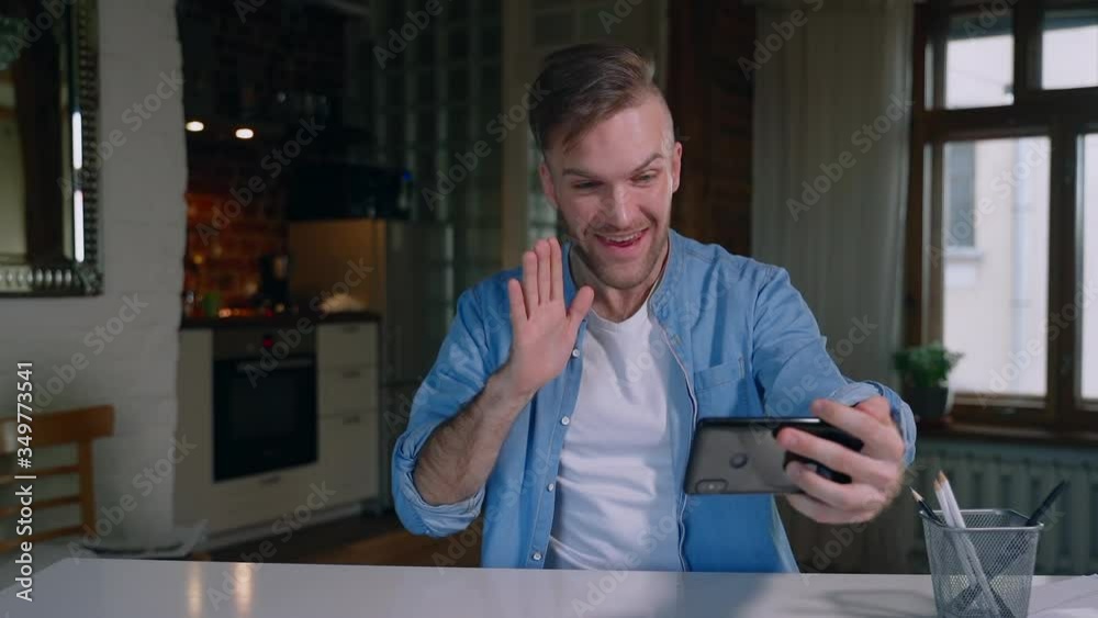 Young man communicates in video chat using smartphone during self-isolation in apartment spbas. Happy bearded guy talks with smile and gestures while holding mobile device in hand. Hipster has good Young man communicates in video chat using smartphone during self-isolation in apartment spbas. Happy bearded guy talks with smile and gestures while holding mobile device in hand. Hipster has good
