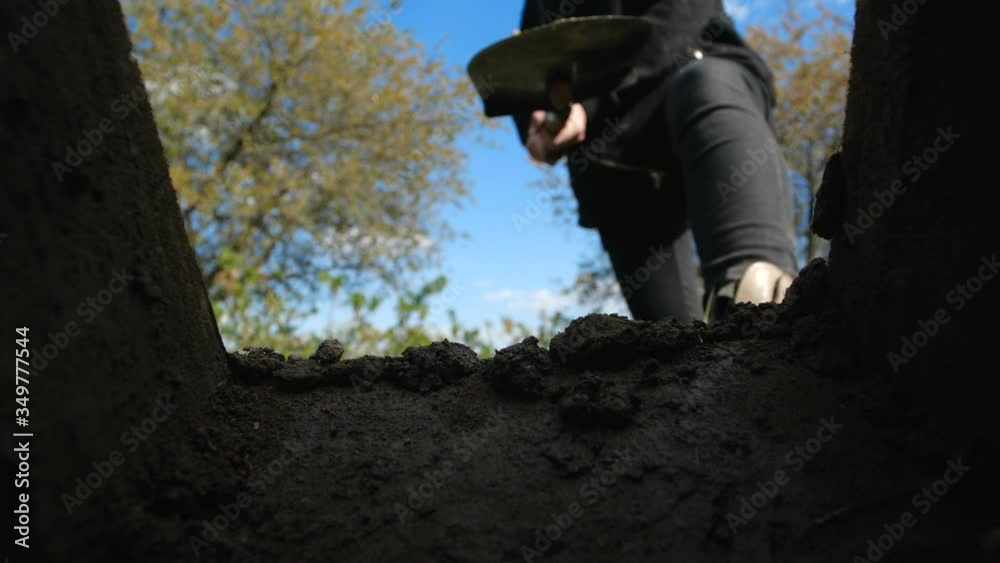 An unrecognizable person in dark clothes is digging a square pit against a blue sky with clouds. Point of view from the grave into which soil is sprinkled from shovel.  Slow motion 200 fps
