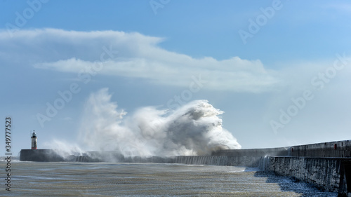 Fotografie Massive waves crash over harbour wall onto lighthouse during huge storm on Engli