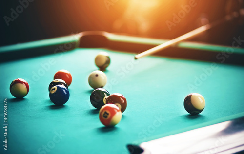 On a green billiard table are billiard balls, and a man with a cue in his hands, illuminated by a bright light, aims at one of the balls.