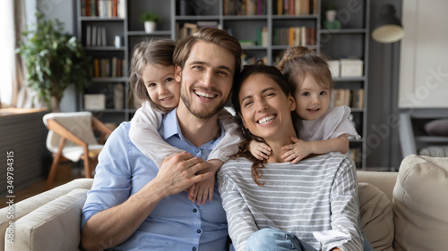 Portrait of young Caucasian family with small daughters pose relax on sofa in living room, smiling little girls kids hug embrace parents, show love and gratitude, rest on couch at home together