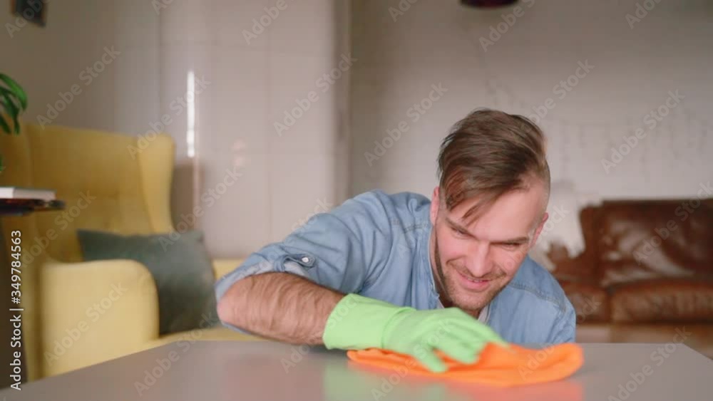 Bearded guy hipster is cleaning table and staying at home during quarantine spbas. Young american man rubs wooden surface with rag and smiles while spending day in apartment. Person is doing household Bearded guy hipster is cleaning table and staying at home during quarantine spbas. Young american man rubs wooden surface with rag and smiles while spending day in apartment. Person is doing household