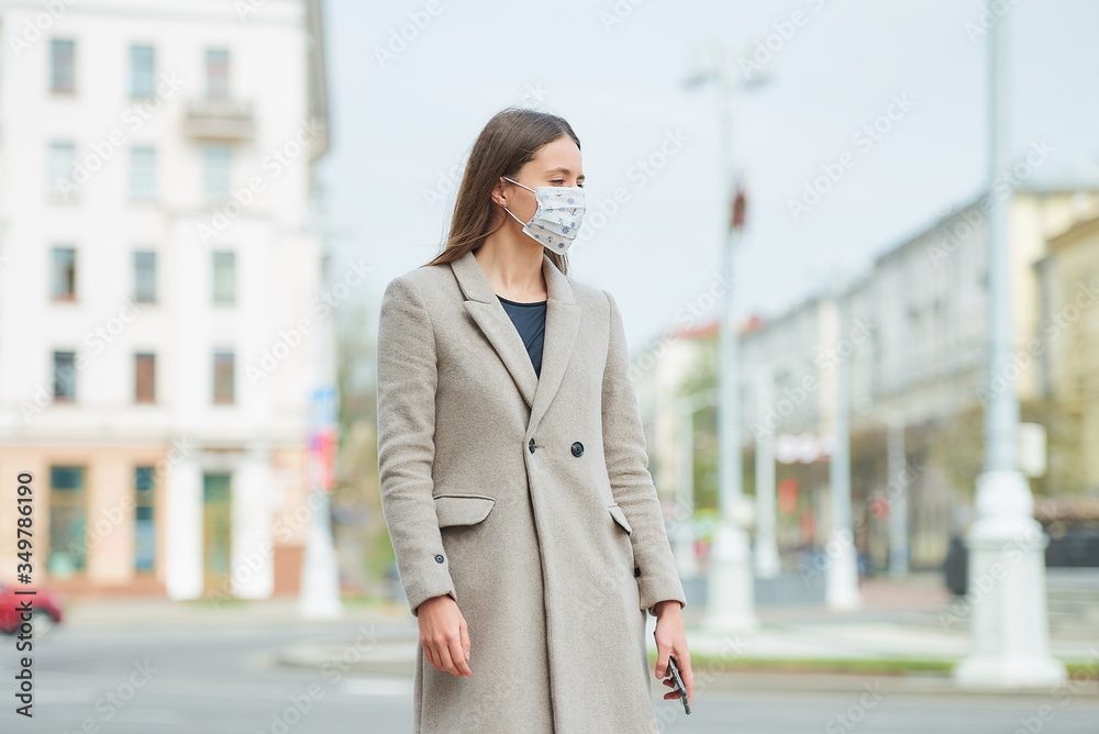 A brunette girl with long hair in a medical face mask to avoid the spread coronavirus holds a smartphone in the street. A woman in a face mask against COVID-19 wears a coat looks away in the city.