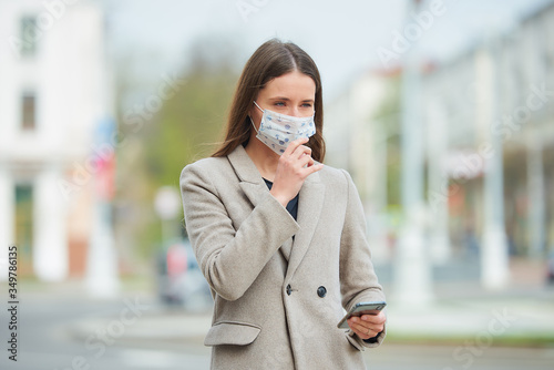 A girl with long hair in a medical face mask to avoid the spread coronavirus uses a smartphone in the street. A woman wears a coat checking a face mask against COVID-19 in the center of the city.