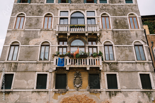 Brown brick stone wall, house facade in Italy, Venice, with old classic Venetian windows with a stone balcony, with a bas-relief in the center and a blue EU flag. © Nadtochiy