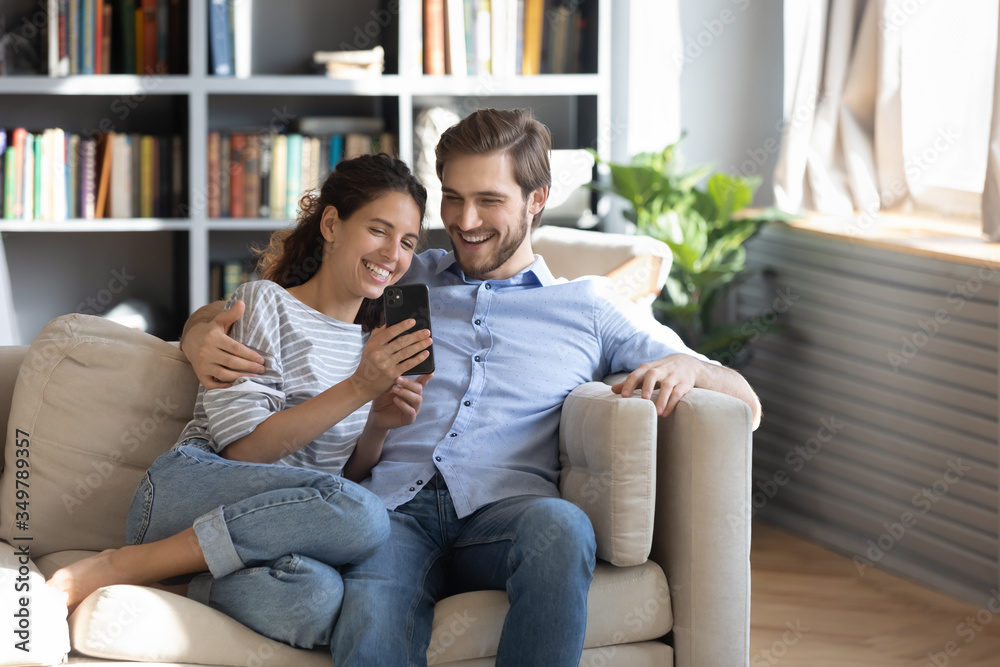 Smiling young couple sit relax on couch in living room have fun using smartphone together, happy Caucasian man and woman rest on sofa watch video on cellphone, make self-portrait picture on cell