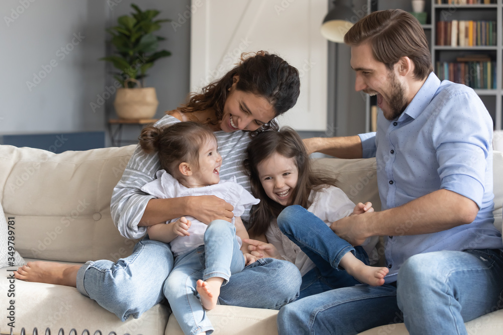 Overjoyed Caucasian family with little preschooler daughters sit relax on couch in living room playing tickling, happy young parents with small kids rest on sofa at home, have fun on weekend together