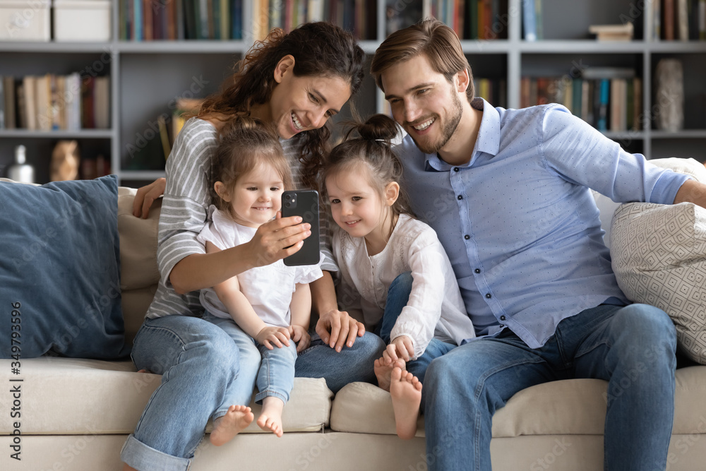 Smiling young Caucasian family with little daughters sit on couch in living room male selfie on smartphone together, happy parents with small children pose for self-portrait picture on cellphone