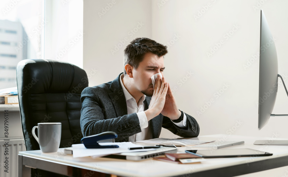 A young businessman or office worker sneezes, coughs, blows his nose at the workplace. Overworked worker got colds and flu. High temperature with ARVI or SARS. Epidemic of influenza or coronavirus. A young businessman or office worker sneezes, coughs, blows his nose at the workplace. Overworked worker got colds and flu. High temperature with ARVI or SARS. Epidemic of influenza or coronavirus.
