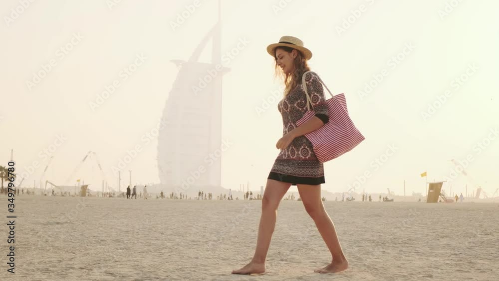 Young beautiful cute woman tourist walking sunset along shore Persian Gulf, sandy plage, coastline Dubai Beach. Girls blonde long hair. Backdrop summer sky, foggy image modern skyscraper. UAE 2020 4k
