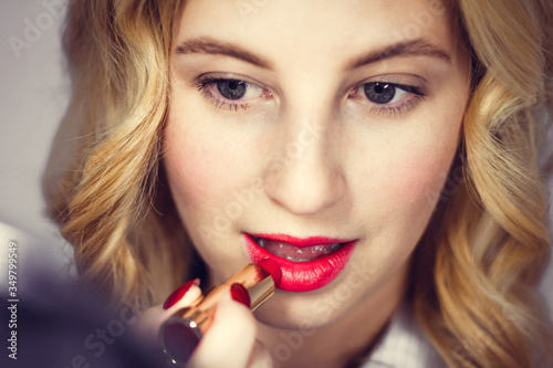 Attractive blonde paints her lips with bright red lipstick. Sexy cute girl with white skin does makeup with mirror. Decorative cosmetics. Beauty industry