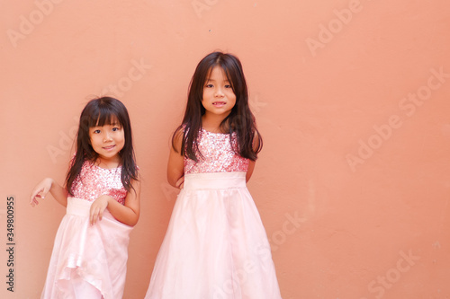 The photo of two lovely little Asian girls stand in front of the orange brown blank wall background.