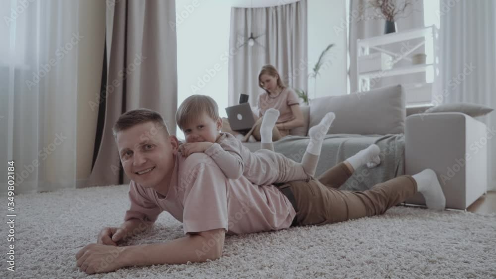 Young dad spends time with his little son, they lie together on the floor and smile, and mom in the background sits on the couch with a laptop and shopping online Young dad spends time with his little son, they lie together on the floor and smile, and mom in the background sits on the couch with a laptop and shopping online
