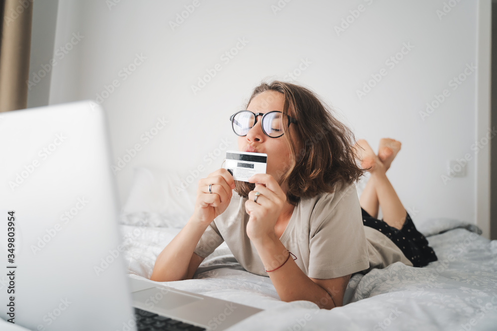 Beautiful happy young curly woman lies on a bed in front of a laptop and kisses a credit card 