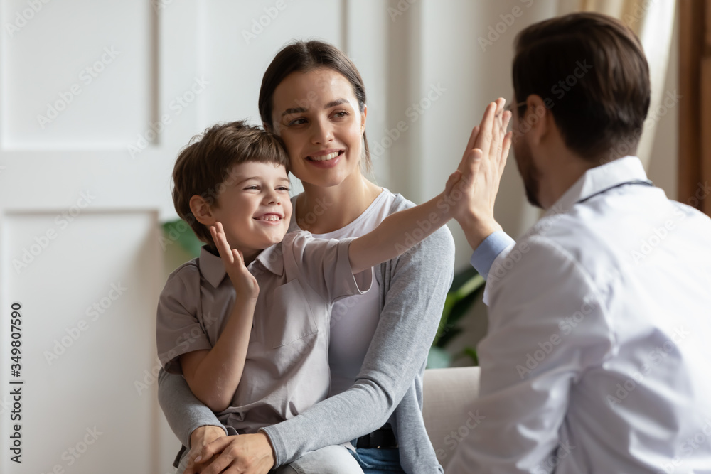 Overjoyed little boy patient have fun give high five to male doctor at consultation in clinic with mom, happy small child greeting with man pediatrician at checkup in hospital, healthcare concept