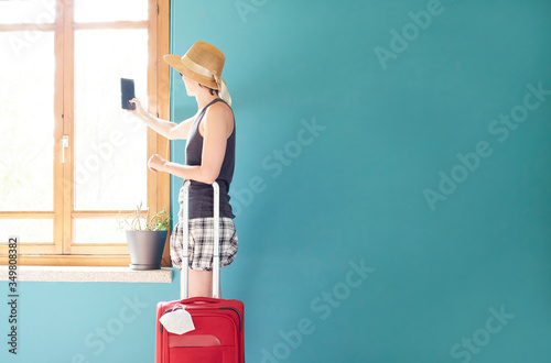 Woman in skirt, with straw hat and striped shirt using her mobile prepared with a red suitcase and her face mask for her vacation in Covid's time.
